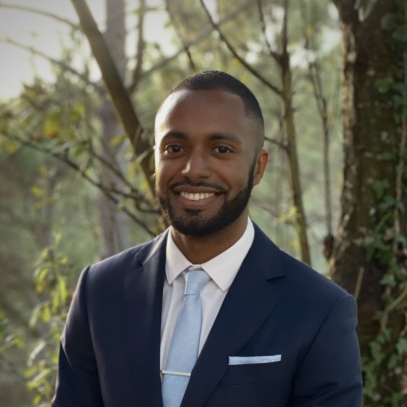 Man wearing a navy blue suit with a light blue tie and matching pocket square standing outdoors among trees.