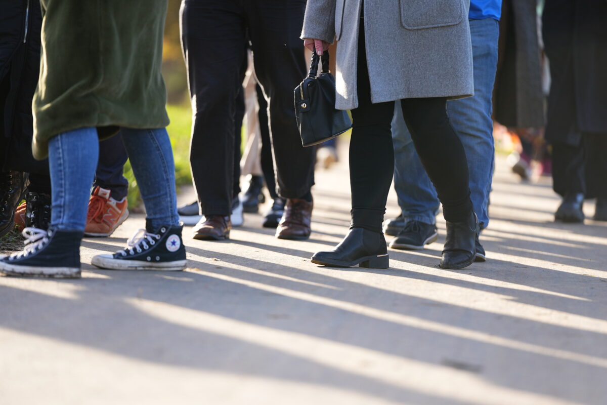 A group of people walking outdoors on a sunny day. The focus is on their legs and footwear, showing a mix of boots, trainers, and shoes as they walk together along a paved path. Shadows stretch across the ground.