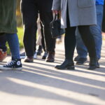 A group of people walking outdoors on a sunny day. The focus is on their legs and footwear, showing a mix of boots, trainers, and shoes as they walk together along a paved path. Shadows stretch across the ground.