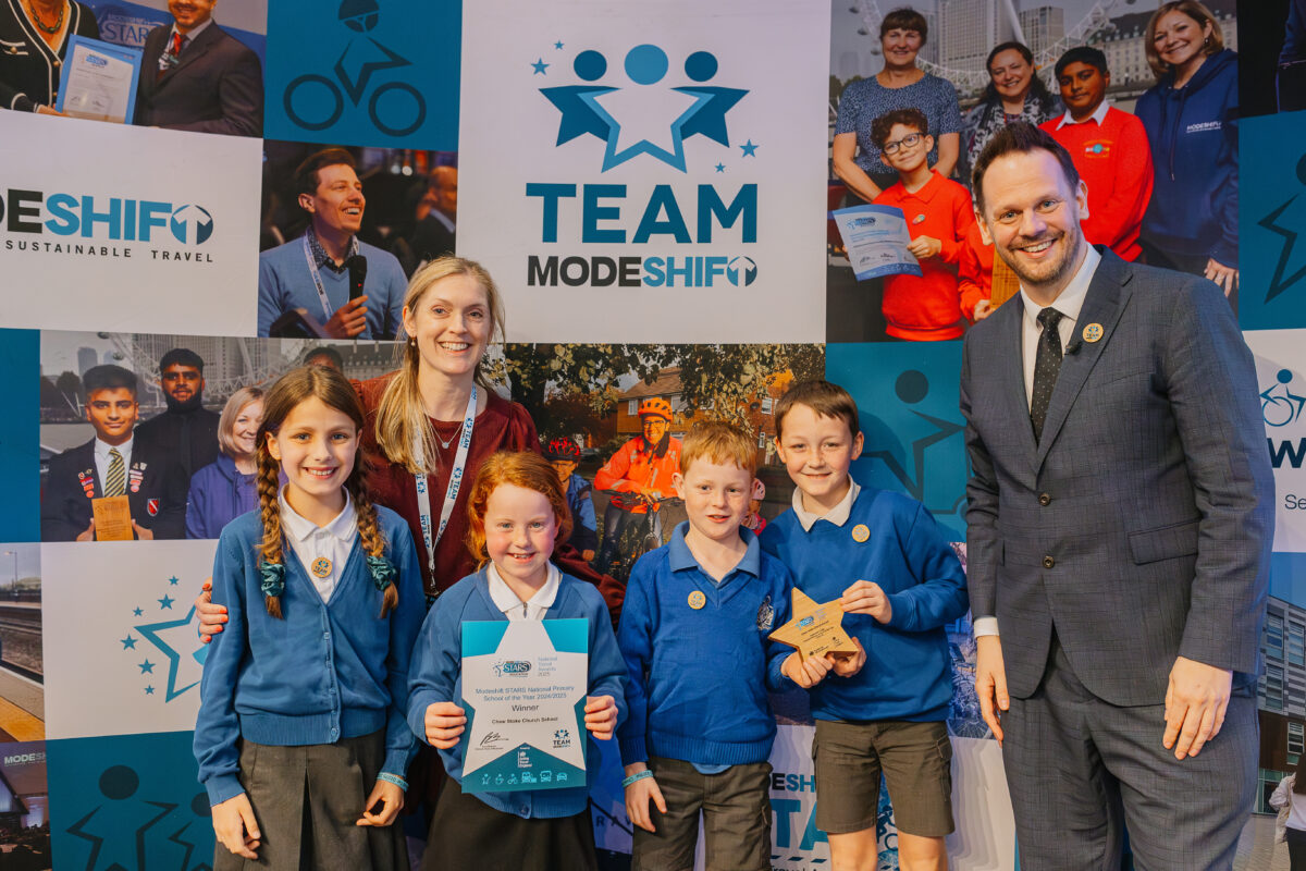A group of four school children and two adults smile at the camera. Two of the children dressed in school uniform hold up a star shaped award and certificate.