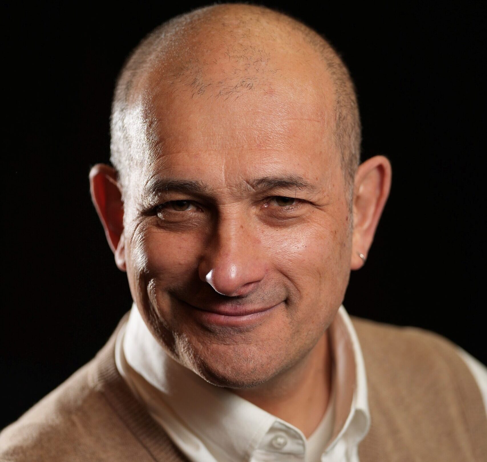 Man wearing a white collared shirt under a brown sleeveless sweater against a black background.