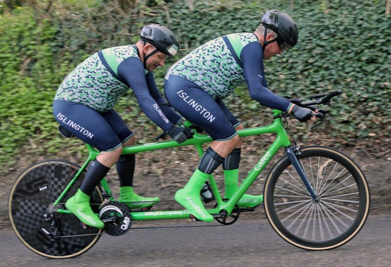 Two cyclists in matching Islington gear ride a bright green tandem bike along a paved path with greenery in the background.