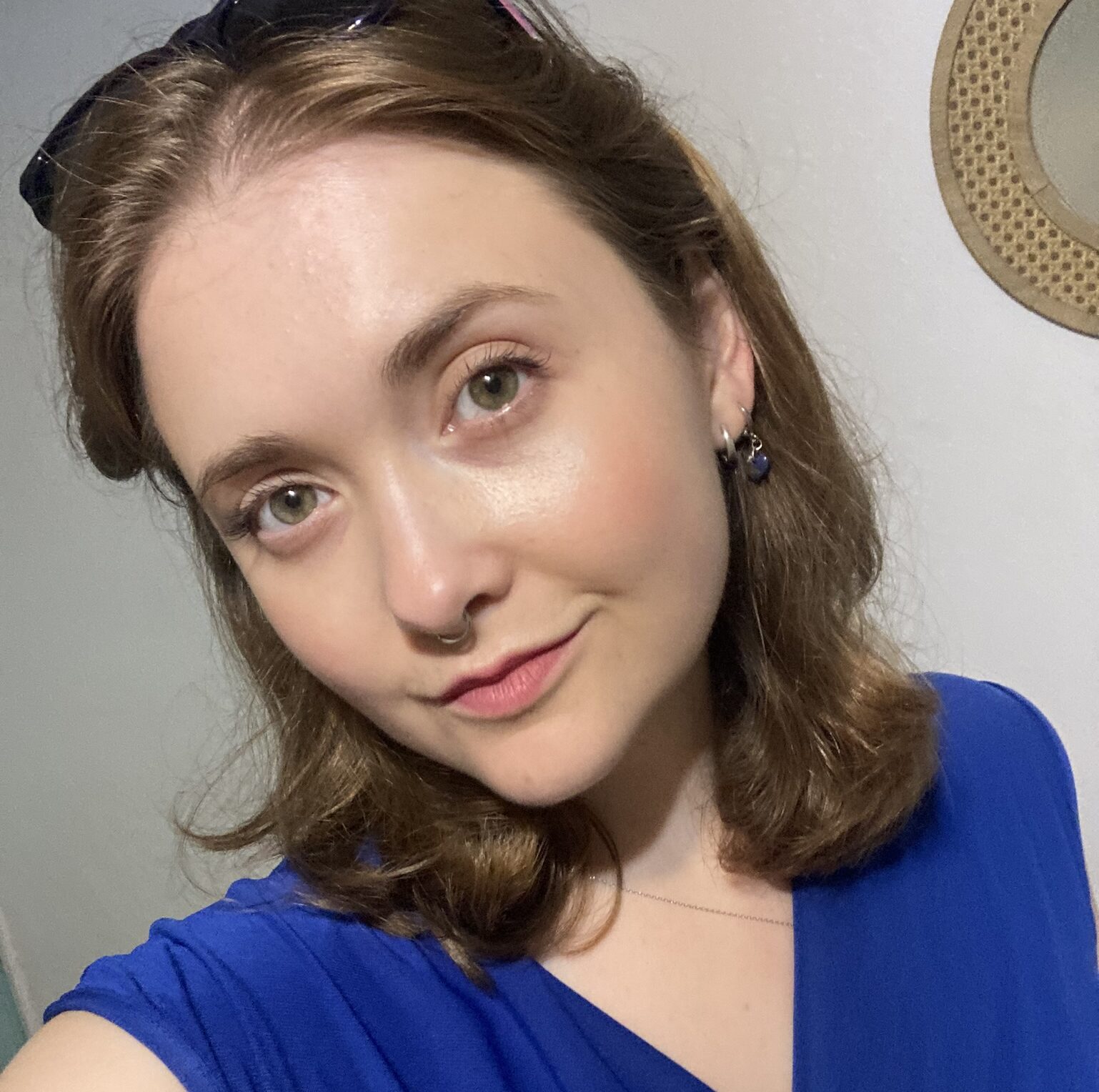 Woman wearing a vibrant blue top with short sleeves and a delicate necklace, standing indoors near a wall with a decorative round mirror.