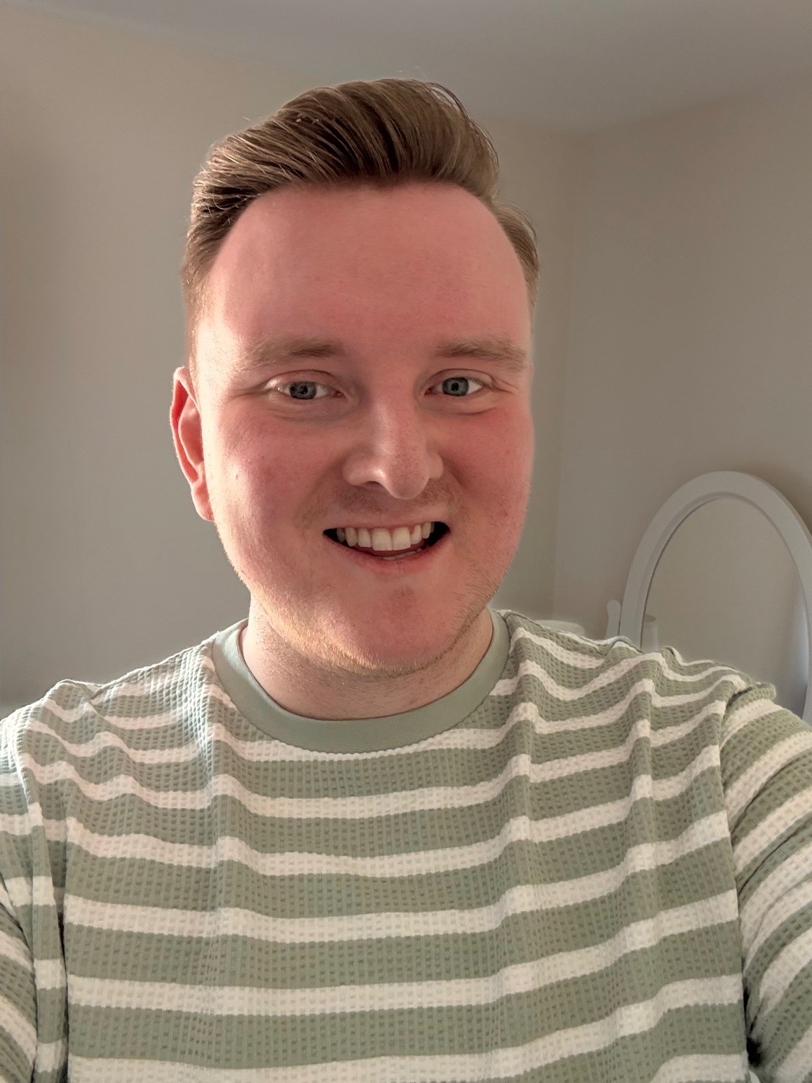 A man smiling indoors, wearing a light green and white striped shirt, standing in front of a cream wall with a mirror in the background.