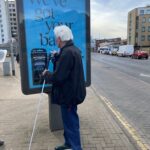 Elderly man with white hair and a walking cane reading a blue advertisement for Wilkinson Sword Hydro razors on a city sidewalk.