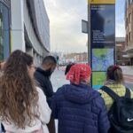 Group of people standing on a city sidewalk near an Eden Street and Brook Street directional sign with a map and local landmarks listed.