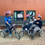 Three people on adaptive tricycles holding small Union Jack flags outside a brick building with a colorful sign in the window.