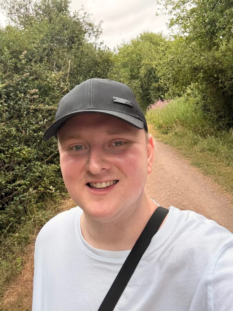 A man smiling outdoors on a tree-lined path, wearing a black cap, a white T-shirt, and a crossbody strap.