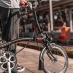 Person standing next to a black Brompton folding bike on a train station platform, holding the handlebars ready to ride.