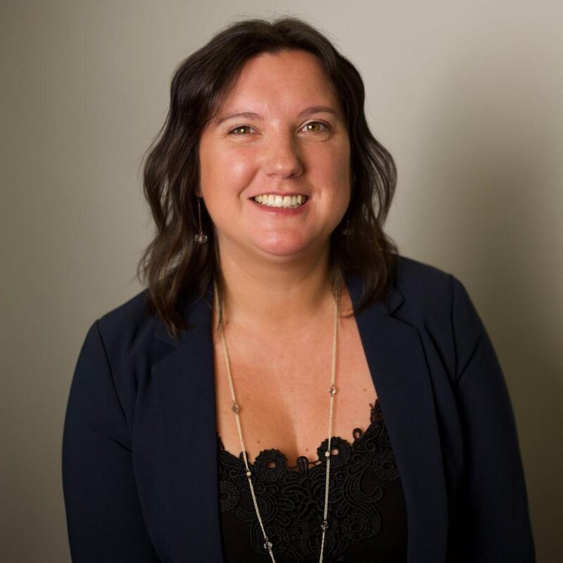 Woman wearing a navy blazer over a black lace top with a long delicate necklace against a plain beige background.