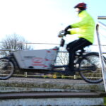 Person wearing a bright yellow jacket and helmet riding a cargo bike over a small bridge with metal railings on a sunny day.