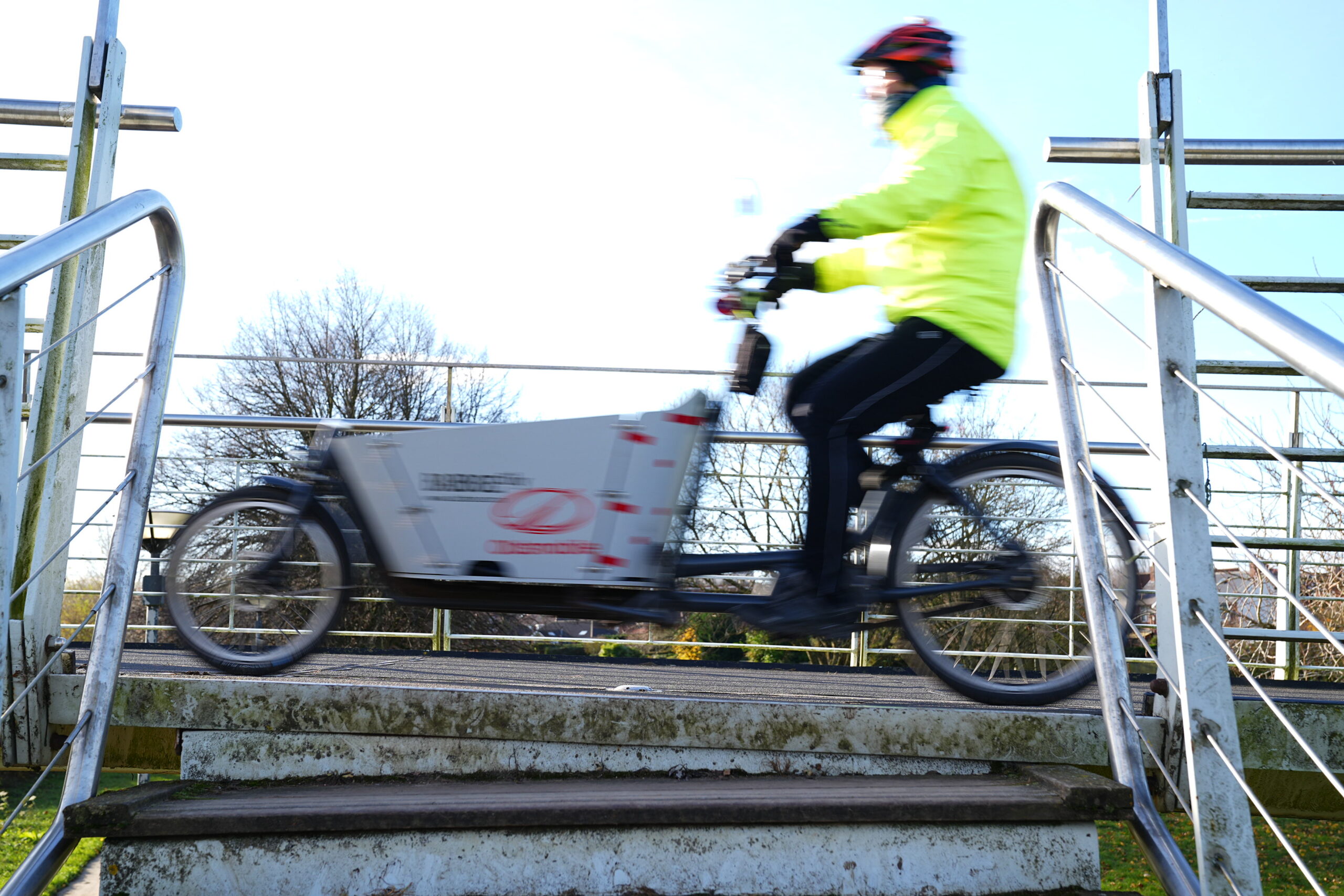 Person wearing a bright yellow jacket and helmet riding a cargo bike over a small bridge with metal railings on a sunny day.
