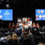 A large event. People are seated around round tables looking toward a large stage with two screens suspended either side of a speaker stood at a lectern in the centre.