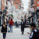 Image credit Active Travel England. A busy high street with people walking. Shops and restaurants either side of a pedestrianised street.