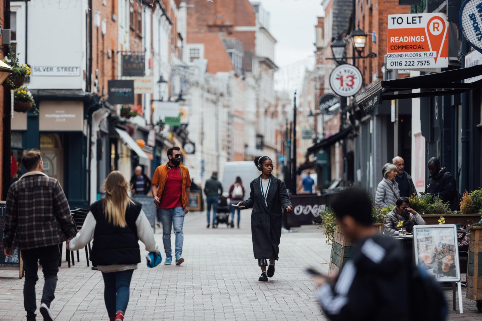Image credit Active Travel England. A busy high street with people walking. Shops and restaurants either side of a pedestrianised street.