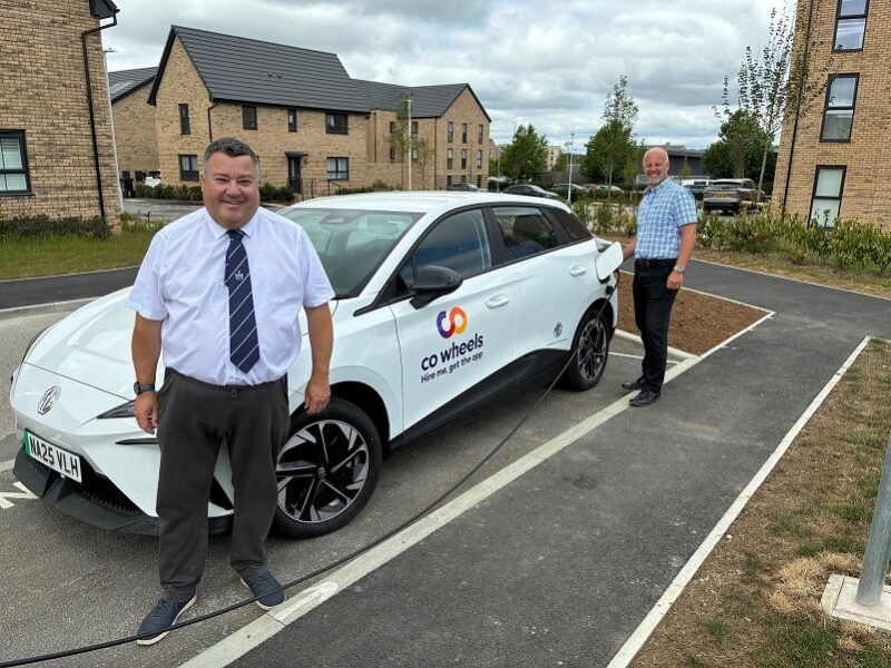 Two men stand beside a white CO Wheels electric car parked in a residential neighborhood with modern brick houses and cloudy skies.