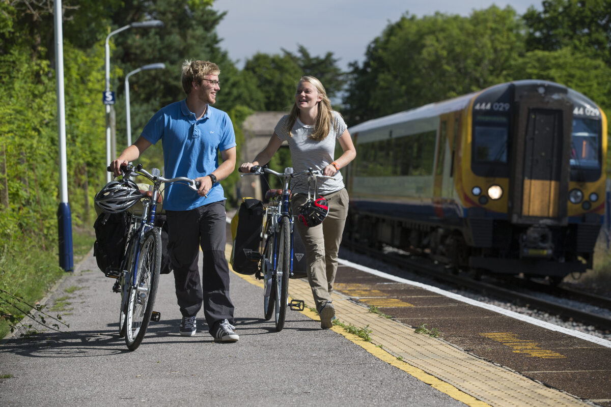 A man and a woman push their bikes side by side, a train platform with train in the background. Image credit New Forest National Park.