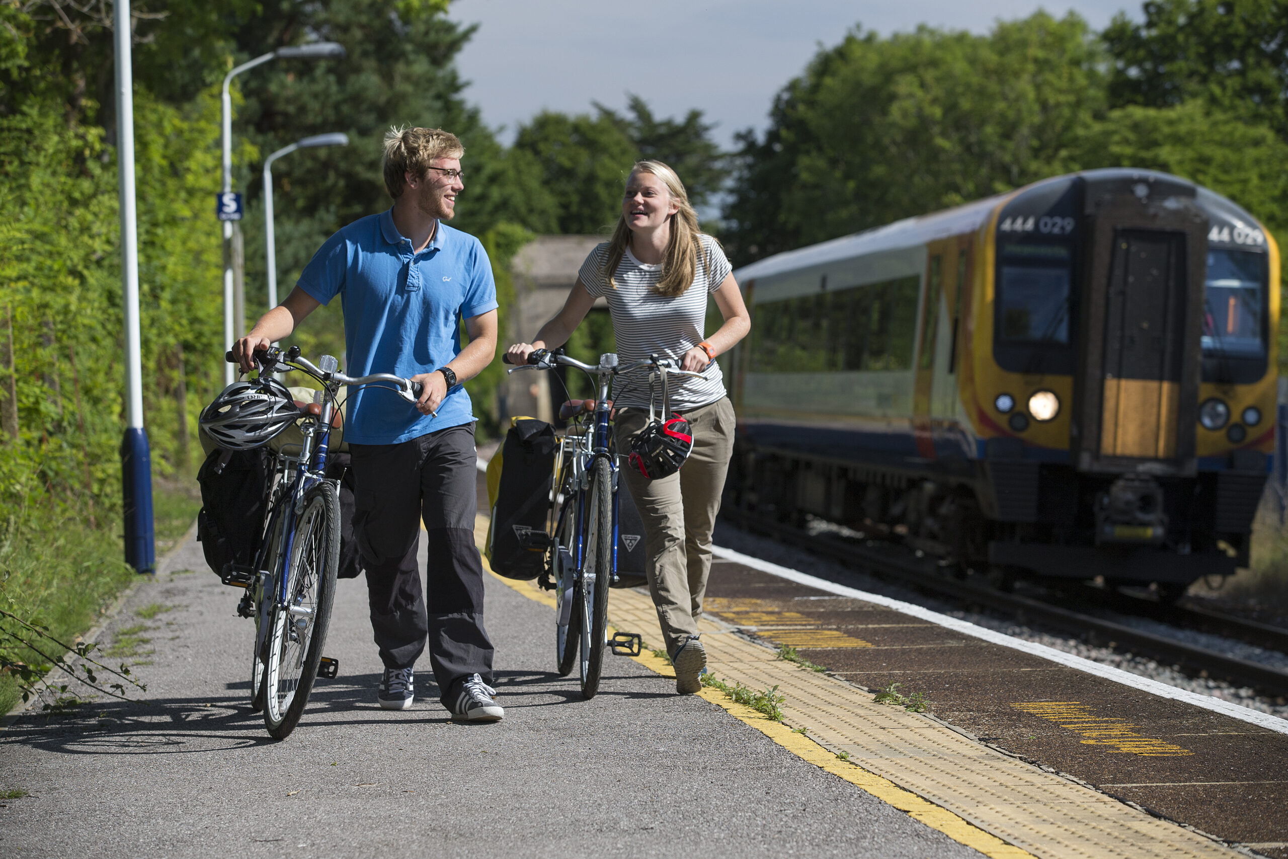 A man and a woman push their bikes side by side, a train platform with train in the background. Image credit New Forest National Park.