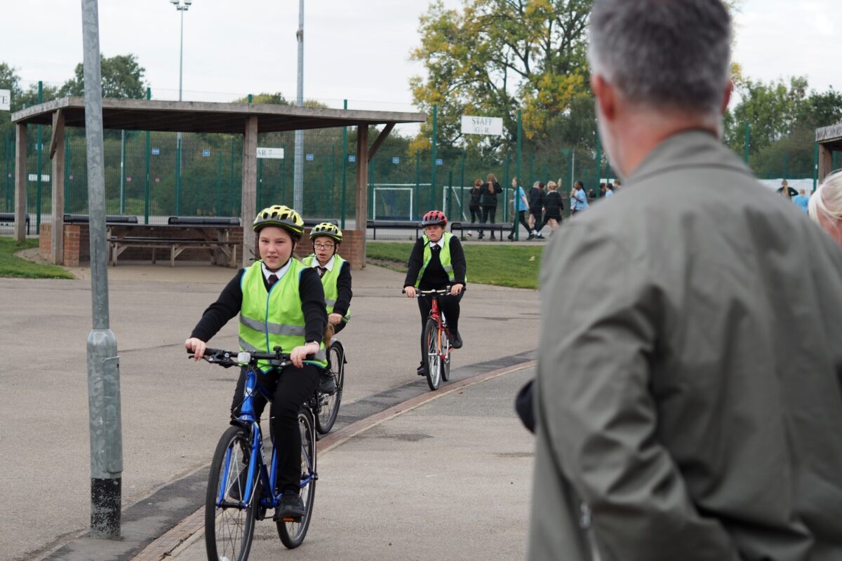 Three school pupils wearing helmets and high-visibility vests ride bicycles on a school playground while an adult looks on. Other students are walking in the background near sports courts and fencing.