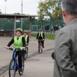 Three school pupils wearing helmets and high-visibility vests ride bicycles on a school playground while an adult looks on. Other students are walking in the background near sports courts and fencing.