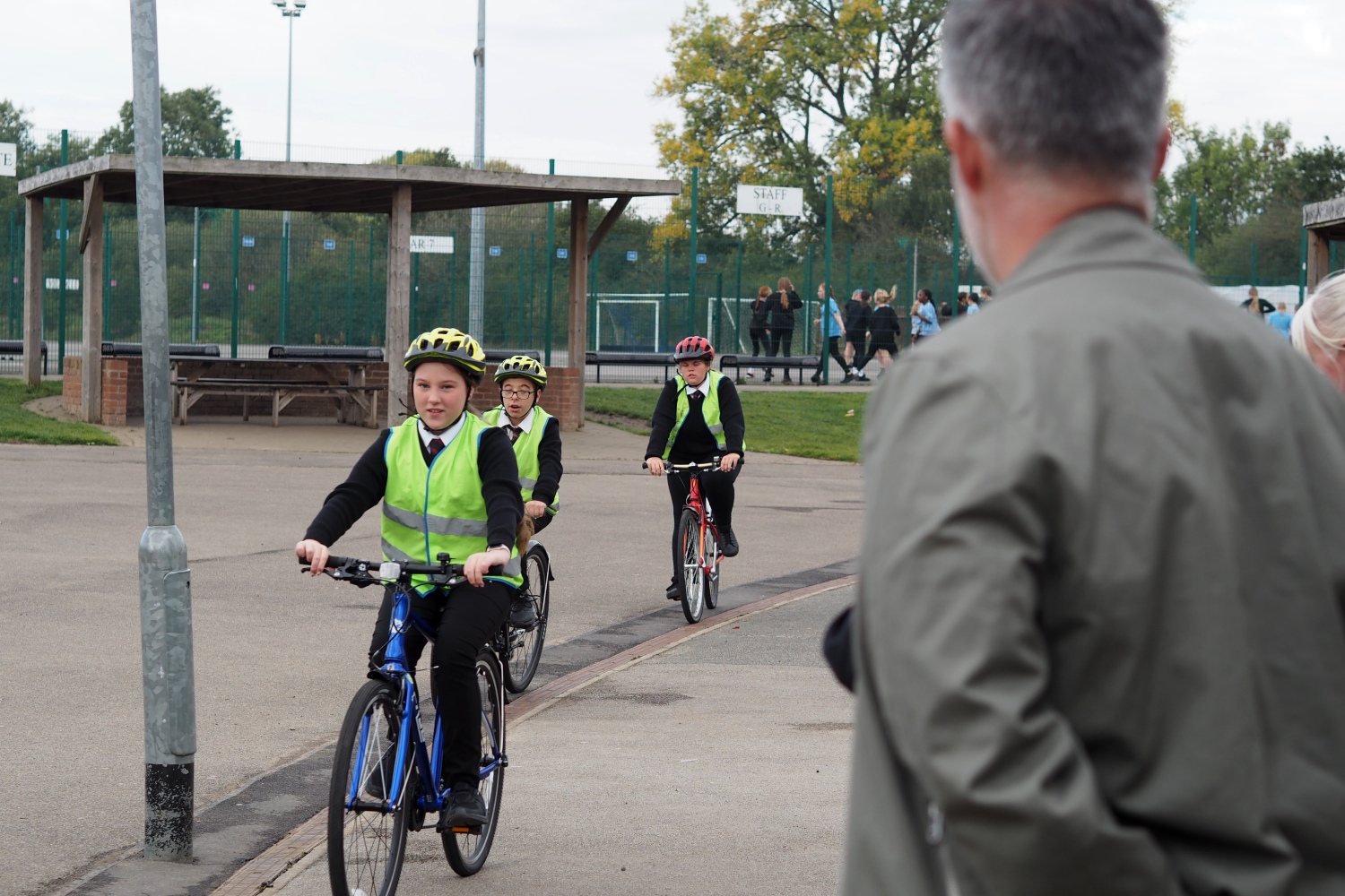 Three school pupils wearing helmets and high-visibility vests ride bicycles on a school playground while an adult looks on. Other students are walking in the background near sports courts and fencing.