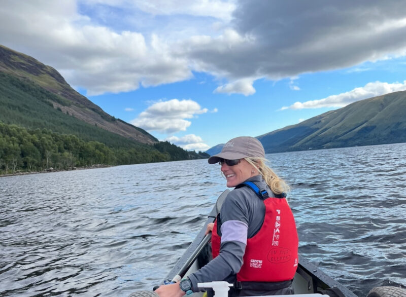 Person wearing a red life jacket paddling a canoe on a calm lake surrounded by green hills under a partly cloudy sky.