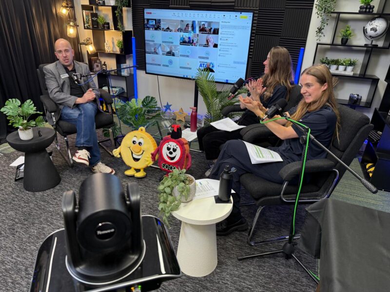 Three people sit in a studio setting during a live or virtual event, speaking into microphones and engaging with an online audience shown on a large screen behind them. Two plush mascots, plants, and studio lights decorate the space, creating a friendly and professional atmosphere.