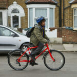 Child cycles down a residential street on a red bike. Wears green winter coat and blue helmet.