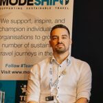 Man in a white shirt sitting at a table with a MODESHIFT banner promoting sustainable travel behind him.