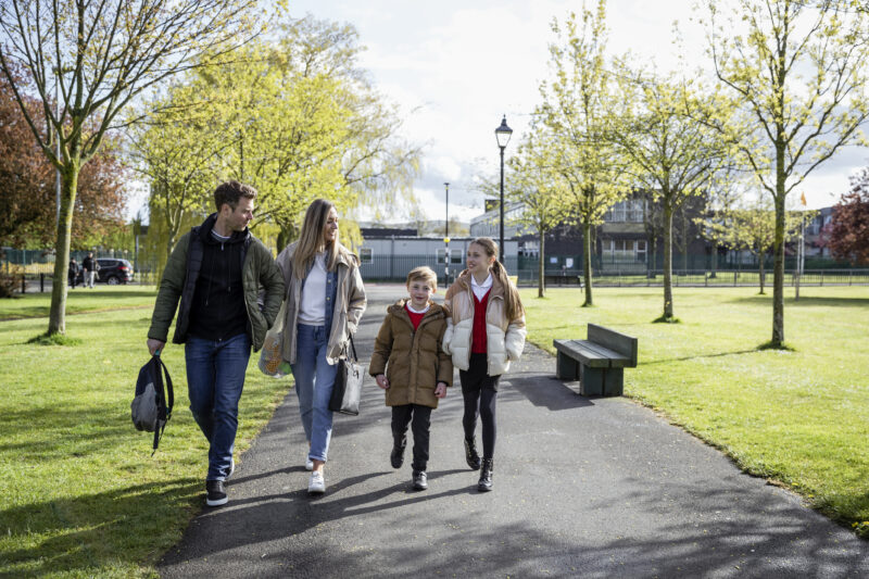 A family of four walks together along a park pathway on a bright day. Two adults and two children, dressed warmly in jackets, appear to be walking to or from school, chatting and smiling as they go. Trees with spring leaves and a school building are visible in the background.
