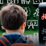 Child with backpack pressing pedestrian crossing button showing red bicycle and walking figure signals.