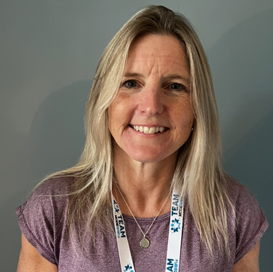 Woman with long blonde hair smiles at the camera. She wears a lilac t-shirt and a Modeshift branded lanyard around her neck.