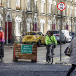 Two cyclists ride on a street with a sign indicating the road is open to pedestrians, wheelchair users, and cyclists only.