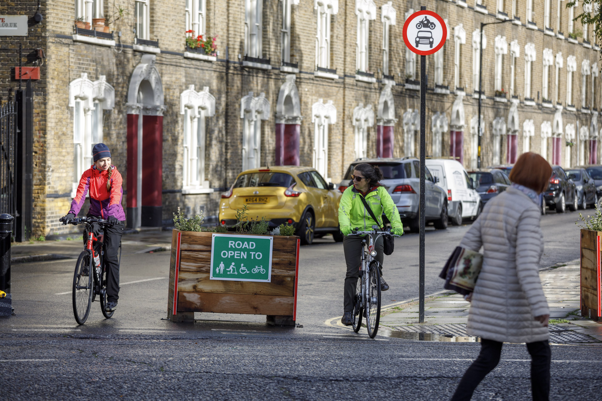 Two cyclists ride on a street with a sign indicating the road is open to pedestrians, wheelchair users, and cyclists only.