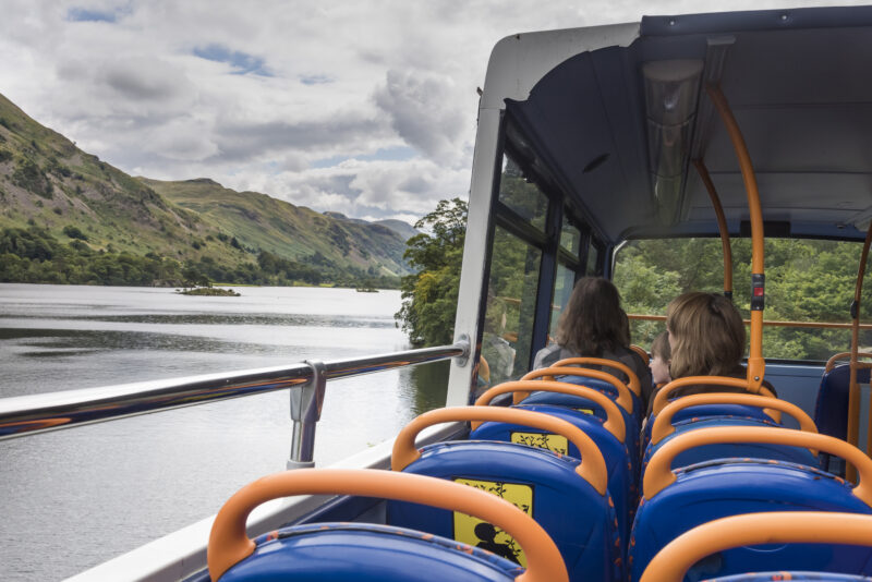 Passengers on an open top bus pass an expansive lake, mountains and cloudy sky in the background. Image Credit: Go Lakes