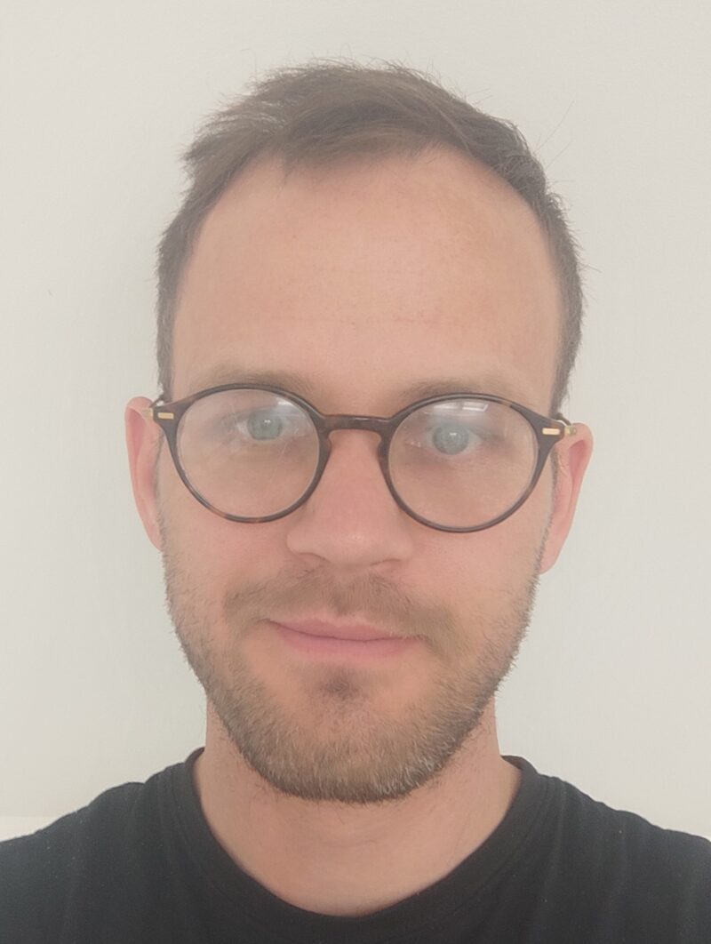 A man smiles at the camera, head and shoulder shot. He wears dark rimmed glasses and a black top.