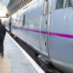 A railway platform. A train is stationery with people walking on the platform, they are blurred as is suspended in movement.