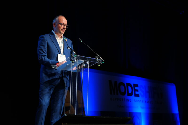Man stands on a stage at a lectern addressing the audience. Modeshift branded stage set in the background.