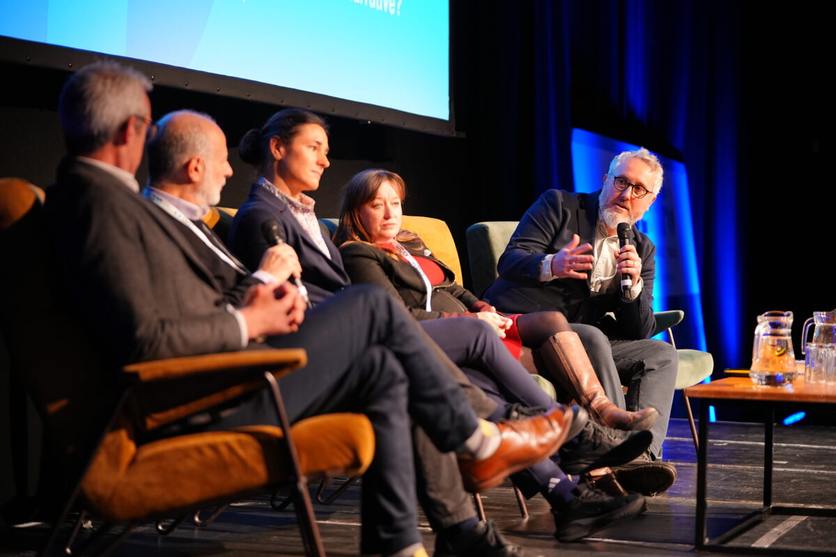 Five people are seated on a stage, a man on the far right holds a microphone and addresses the panel of people.