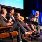 Five people are seated on a stage, a man on the far right holds a microphone and addresses the panel of people.