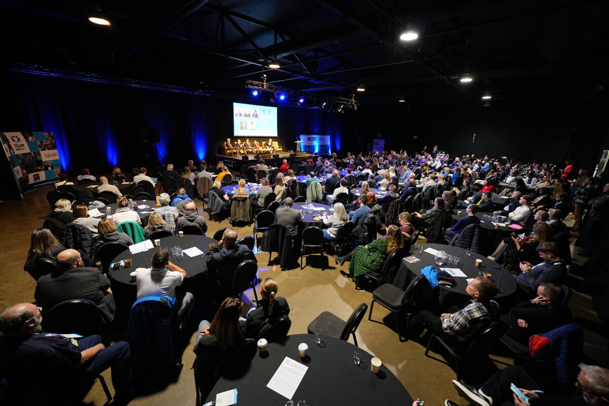 The National Modeshift Convention delegates. Large room with people seated around round tables facing a large screen and stage area.