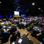 The National Modeshift Convention delegates. Large room with people seated around round tables facing a large screen and stage area.