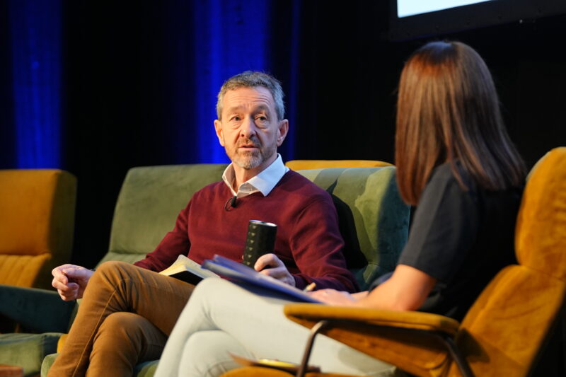Chris Boardman seated speaking onstage, woman sits next to him with notes on her lap.