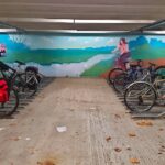 Cycle parking, in the background is a painted mural showing a colourful landscape with cyclist next to a stream.