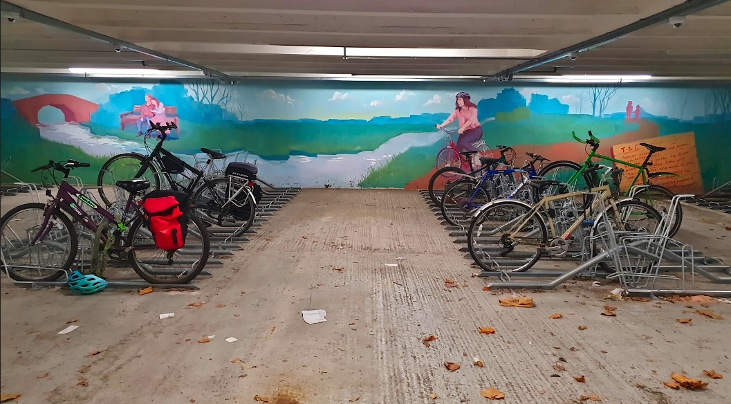 Cycle parking, in the background is a painted mural showing a colourful landscape with cyclist next to a stream.