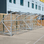 Empty metal bike shelter with transparent roof panels set on a paved area outside a modern building with multiple windows.