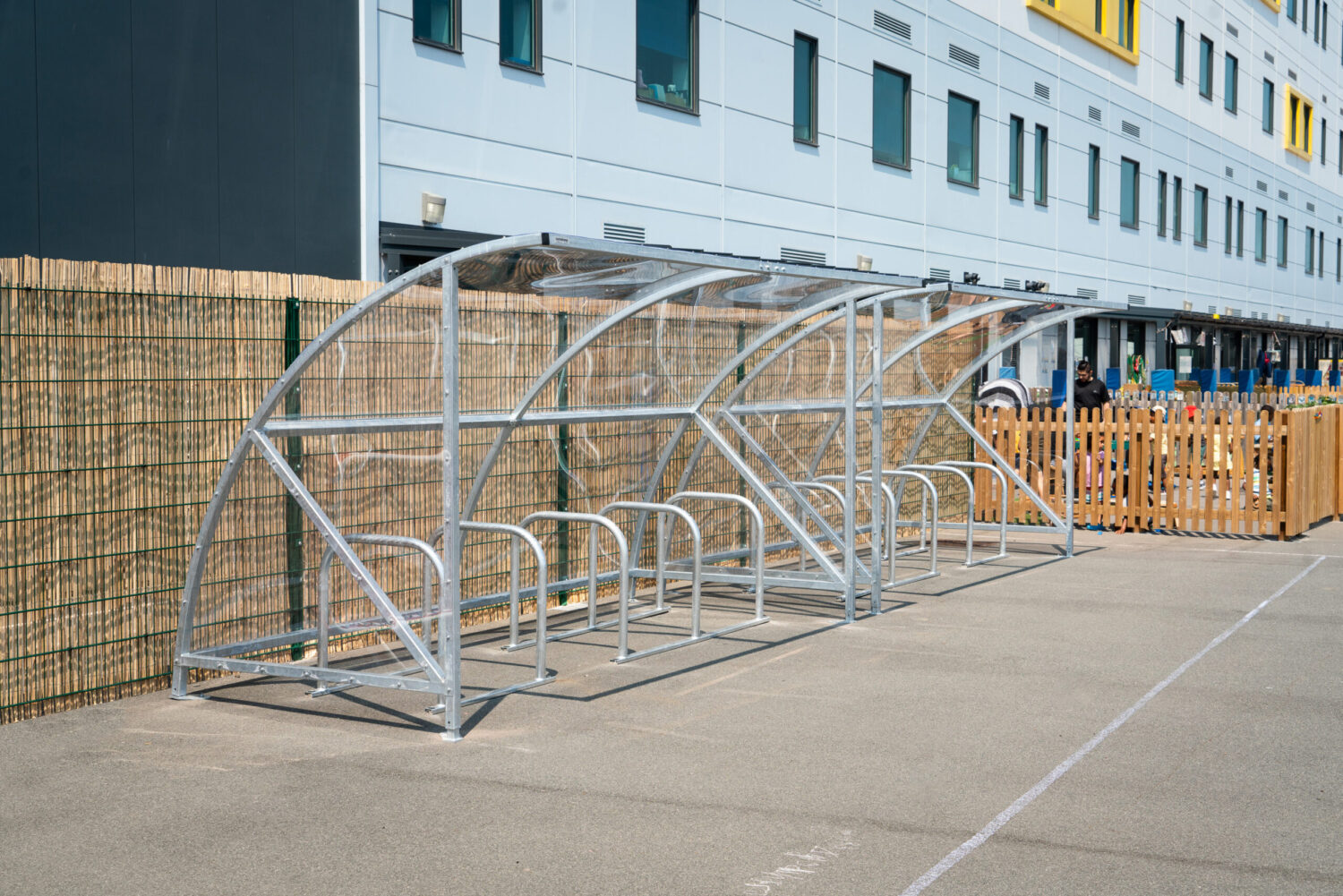 Empty metal bike shelter with transparent roof panels set on a paved area outside a modern building with multiple windows.