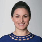 Woman wearing a blue top with a patterned neckline against a plain light gray background.