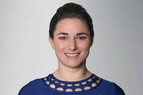 Woman wearing a blue top with a patterned neckline against a plain light gray background.
