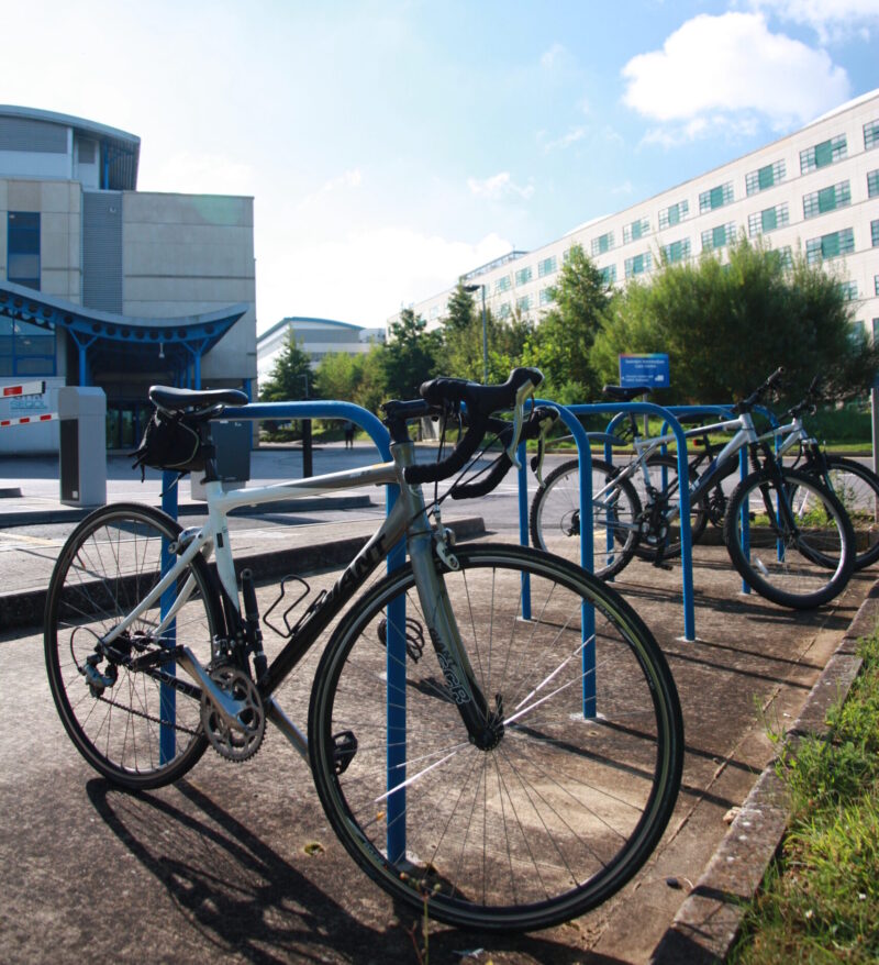Cycles are parked in the foreground. Buildings in the background and a bright blue sky.